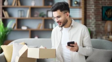 Neşeli, gülümseyen genç, yetişkin bir müşteri paketini evde açıyor. Mutlu erkek oturma odasındaki kanepede oturan akıllı telefondan iyi eleştiriler yazar. E-ticaretin tatmin olmuş müşterisi. Ekspres dağıtım hizmetleri