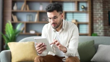 Evdeki oturma odasındaki kanepede dijital tablet kullanan yakışıklı adam. Gözlüklü, gülümseyen sakallı bir erkek sosyal ağlara takılır, çevrimiçi bir mağazada alışveriş yapar, sohbet eder, yazar veya mesajlar okur.