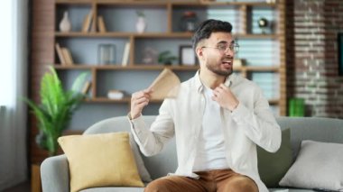 Yakışıklı sakallı adam evdeki oturma odasında kanepede otururken çok seksi. Genç yetişkin erkek yelpaze sallıyor, rahatsız ve terliyor, çünkü evdeki klima bozuk.