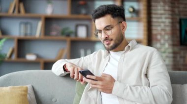 Evdeki oturma odasında koltukta oturan cep telefonu kullanan yakışıklı sakallı adam. Erkek sosyal ağlara takılır, çevrimiçi bir mağazada alışveriş yapar, arkadaşlarıyla sohbet eder, mektup yazar veya okur. Kapat.