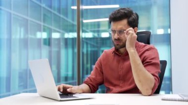 Göz yorgunluğu ve baş ağrısından muzdarip çok çalışan sakallı iş adamı iş yerindeki bir laptopta çalışıyor. Yorgun girişimci gözlüklerini çıkarır ve masajlar burun köprüsünü ovalar.