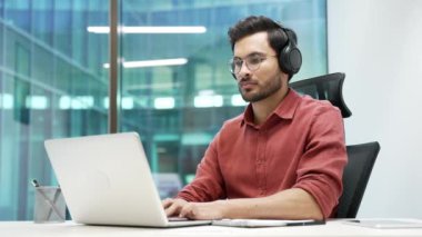 İş yerindeki bir laptopta çalışan, kablosuz kulaklık üzerine çalışan yakışıklı bir bilişim uzmanının portresi. Kameraya bakan başarılı gülümseyen geliştirici ya da kodlayıcı