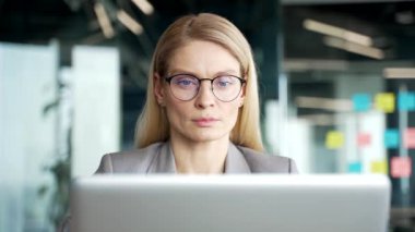 Ciddi düşünceli iş kadını iş yerindeki iş yerinde dizüstü bilgisayarda çalışıyor. Gözlüklü kadın çalışan bir projeyle meşgul. Bankacılık, mesajlaşma ya da sohbet. Ön manzara. Kapat.