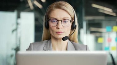 Kablosuz kulaklık takan kendine güvenen kadın müşteri temsilcisi bilgisayarla iş yerinde otururken görüntülü konuşma yapıyor. Çağrı merkezi temsilcisi müşteriye tavsiyede bulunur. Kapatın. Ön görünüm