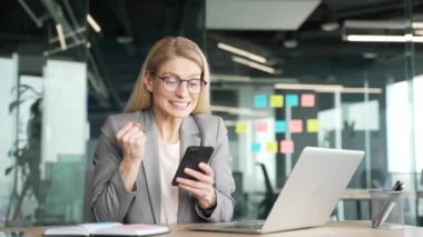 İş yerinde masa başında otururken ceketli mutlu iş kadını cep telefonundan harika haberler okuyor. Neşeli kadın başarıyı kutluyor, gülümsüyor, akıllı telefondaki olumlu mesajla seviniyor.