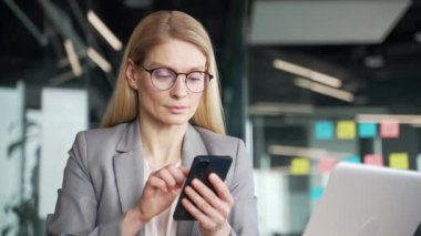 Ceketli mutlu iş kadını iş yerinde otururken cep telefonundan harika haberler okuyor. Gülümseyen mutlu kadın başarıyı kutlar, gülümser, olumlu mesajla sevinir. Kapat.