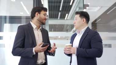 Takım elbiseli iki profesyonel iş adamı modern ofiste akıllı telefon ve kahve ile gündelik tartışmalara giriyorlar. Meslektaşlar dostça sohbet eder. Takım çalışması, iletişim, iş birliği