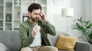 Evdeki oturma odasında kanepede otururken göz yorgunluğu ve baş ağrısı çeken yakışıklı bir adam. Aşırı yorgun yetişkin sakallı erkek gözlüğünü çıkarır ve eliyle burnunun köprüsüne masaj yapar.