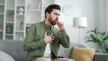 Evdeki oturma odasında kanepede otururken öksüren yakışıklı adam. Soğuk algınlığı ya da grip sakallı bir erkek elini göğsüne dayayıp kendini kötü hisseder. Bronşit ya da akciğerlerinde iltihap var.