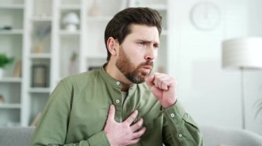 Evdeki oturma odasında kanepede oturan yakışıklı adam öksürüyor. Soğuk algınlığı ya da grip sakallı bir erkek elini göğsüne dayayıp kendini kötü hisseder. Bronşit ya da akciğerlerinde iltihap var. Kapat.