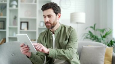 Evdeki oturma odasındaki koltukta dijital tablet kullanan mutlu yakışıklı adam. Gülümseyen sakallı bir erkek sosyal ağlara takılır, çevrimiçi bir mağazadan alışveriş yapar, sohbet eder, yazar ya da mesajları okur.