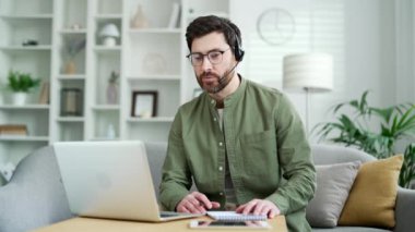 Kulaklıklı, kendine güvenen, sakallı, yakışıklı bir öğretmen. Ev ofisinde otururken bilgisayarla konuşuyor. Koç ya da öğretmen çevrimiçi bir eğitim, seminer, sınıf, kurs ya da webinar düzenler.