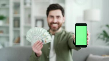 Elinde para tutan ve cep telefonu uygulamasını boş yeşil ekranlı gösteren memnun bir adam. Gülümseyen iş adamı para kazanıyor evdeki kanepede oturan dolarların hayranı. Renk anahtarı, şablon düzeni