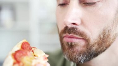 Kapatın. Tatmin olmuş sakallı adam içeride pizza yiyor ve kapalı gözlerle eğleniyor. Neşeli yakışıklı erkek, lezzetli yemeğin tadını çıkarıyor. Eve hızlı bir şekilde yemek teslimatı yapılmasından mutlu.