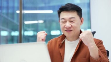 Memnun Asyalı işadamı iş yerindeki bir masada otururken dizüstü bilgisayarından iyi haberler aldı. Gülümseyen mutlu girişimci bilgisayardan olumlu bir mesaj okuyor. Kapat.