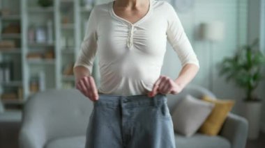Oturma odasında dikilirken bol pantolon giymiş bir kadın önemli miktarda kilo vermiş. Kilo yönetimi, sağlıklı yaşam tarzı ve kişisel fitness konularında başarı kavramı. Kapat.