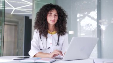 Modern bir hastane kliniğinde dizüstü bilgisayar üzerinde çalışan beyaz önlüklü mutlu bir kadın doktorun portresi. Yetişkin genç kadın tıp doktoru ofisteki iş yerinde kameraya bakıyor.