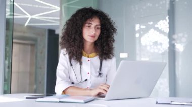 Odaklanmış genç bayan doktor, bir hastane kliniğinde çalışma masasında otururken dizüstü bilgisayar kullanıyor. Esmer bir kadın doktor modern ofisteki bir bilgisayarda çalışıyor.