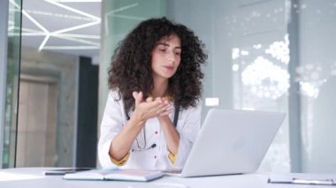 Modern hastane kliniğinde dizüstü bilgisayarla konuşan beyaz önlüklü, gülümseyen genç bayan doktor. Tıp doktoru, ofisteki bir masada oturan bir hastaya danışıyor.