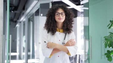 Modern hastane kliniğinde kameraya bakan beyaz önlüklü gülümseyen kadın doktor portresi. Yetişkin pozitif esmer kadın doktor ofiste çapraz kollarla poz veriyor.
