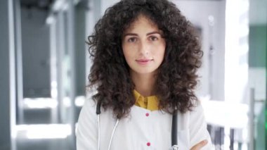 Modern hastane kliniğinde kameraya bakan beyaz önlüklü ciddi bir kadın doktorun portresi. Kendine güvenen genç esmer kadın doktor ofiste poz veriyor. Kapat.