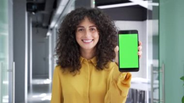 Mutlu genç iş kadını iş yerinde yeşil ekranlı cep telefonunu gösteriyor. Renk anahtarı, reklam için dikey şablon düzeni. Gülümseyen kadın kameraya bakıyor. Elinde akıllı telefon var.