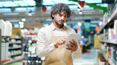 Önlüklü genç personel satıcısı marketteki süpermarkette kameraya bakıyor. Yakışıklı erkek işçi satıcı ya da müdür yiyecek marketi kapalı alanda tablet kullanarak hipermarket gülümsemesi