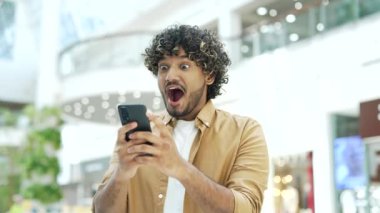 Mutlu genç yakışıklı adam alışveriş merkezindeki akıllı telefona bakarken seviniyor. İnternette indirimli satışlar buluyor. İçerideki zaferi kutlayan sürpriz erkek.