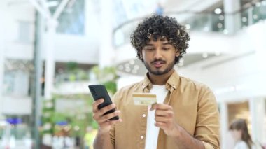 Akıllı telefon ve kredi kartı ile gülümseyen genç ve yakışıklı bir adam alışveriş merkezindeki bir mağazada online ödeme yapıyor, erkek turist akıllı telefon kullanıyor ve ödemeli uygulama satın alıyor.