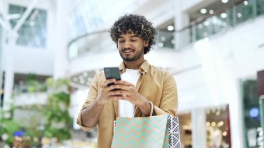 Genç yakışıklı adam alışveriş merkezinde mutlu müşteri duruşu yapıyor ve cep telefonu sohbetleri yapıyor akıllı telefon kullanarak daktilo çalıyor mağazadaki memnun erkek turist kağıt torba hediyesiyle memnun oluyor.