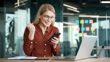İş yerinde otururken cep telefonundan harika haberler okuyan mutlu iş kadını. Neşeli kadın başarıyı kutluyor, gülümsüyor, akıllı telefondaki olumlu mesajla seviniyor.