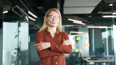 İş yerindeki kendine güvenen bir iş kadınının portresi. Kollarını kavuşturup kameraya bakan gözlüklü ciddi sarışın kadın. Başarılı kadın işçi, modern çalışma alanında poz veriyor.