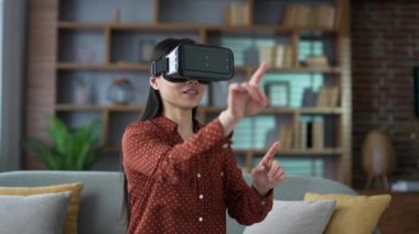 Genç Asyalı kadın sanal gerçeklik simülatöründe sanal gerçeklik gözlüğü kullanarak çalışıyor. Oturma odasındaki kanepede oturuyor. Kadın bilgisayar programlarını kontrol etmek ve sanal sayfaları çevirmek için jestler kullanır