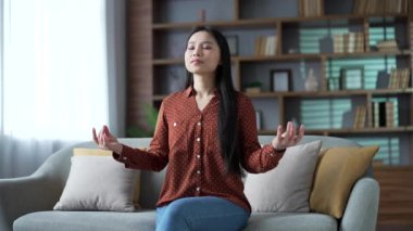 Genç Asyalı kadın, evdeki oturma odasında kanepede otururken gözleri kapalı meditasyon yapıyor. Sakin, mutlu bir dişi yoga yapar, derin derin nefes alır rahat bir atmosferde rahat bir kanepede dinlenir.