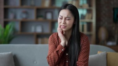 Evdeki oturma odasında kanepede oturan Asyalı kadın diş ağrısı çekiyor. Genç bir dişi, elini yanağında tutuyor ve masaj yapıyor. Keskin bir ağrı hissediyor ve dişçiye görünmeye ihtiyacı var.