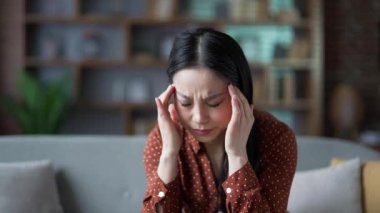 Genç Asyalı kadın evdeki oturma odasında kanepede otururken baş ağrısı çekiyor. Üzgün kadın elleriyle şakaklarına masaj yapıyor. Ağrı hissediyor ve yüksek tansiyonu ya da migreni var. Kapat.