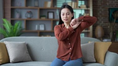 Genç Asyalı kadın evdeki oturma odasında koltukta otururken omzunda keskin bir ağrı hissediyor. Mutsuz dişinin kas zedelenmesi ya da iltihaplanması var. Kız hassas bir noktada kasları ovuyor ve masaj yapıyor.