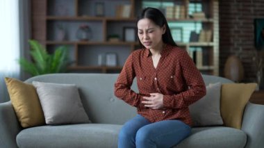 Karın ağrısı çeken genç Asyalı kadın evdeki oturma odasında kanepede oturuyor. Üzgün kadın ellerini karnının üstünde tutuyor. Zehirlenme, kabızlık, şişkinlik ya da adet görme.