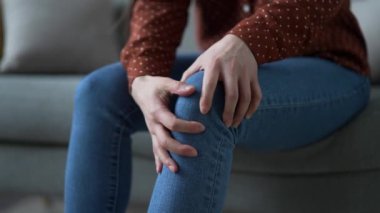Kapatın. Oturma odasındaki kanepede oturan kadın eller diz kaslarına masaj yapıyor. Genç Asyalı kadın diz eklem ağrısı çekiyor. Yürüyemiyor, hastalık, kramp veya romatizma geçiremiyor.
