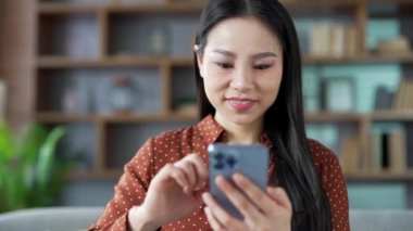 Evdeki oturma odasında cep telefonu kullanan mutlu genç Asyalı kadın gündelik zamanın tadını çıkarıyor. Gülümseyen kadın sosyal medyada geziniyor, sohbet ediyor, mesaj yazıyor ya da okuyor. Kapatın. Ön görünüm