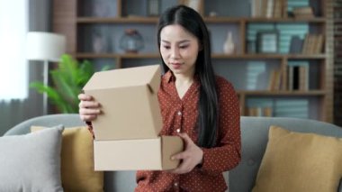 Neşeli genç Asyalı bayan müşteri evde bir paket açıyor. Oturma odasındaki kanepede oturan mutlu kadın alıcı paketleri açıyor. E-ticaretin tatmin olmuş müşterisi. Ekspres dağıtım hizmetleri kavramı