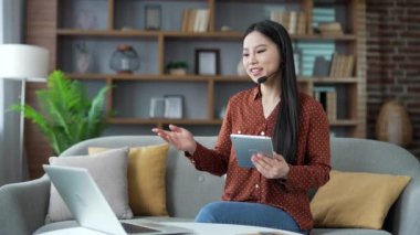 Kablosuz kulaklıklı genç Asyalı iş kadını merkez ofiste bilgisayar ve dijital tablet kullanarak video konferansında konuşuyor. Kendine güvenen bayan koçun çevrimiçi bir eğitimi veya kursu var.