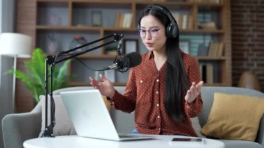Asyalı kendine güvenen genç bir kadın merkez ofiste dizüstü bilgisayar ve mikrofon kullanarak podcast yapıyor. Vlogger etkileyicisi çevrimiçi bir yayın, röportaj veya webinar yönetir, video çağrısı ile uzaktan konuşur