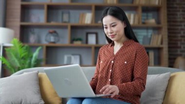 Dizüstü bilgisayarda çalışan, evdeki oturma odasında oturan genç, gülümseyen Asyalı kadının portresi. Dost canlısı kadın kameraya bakıyor. Mutlu pozitif öğrenci bilgisayardaki bir görevle meşgul.
