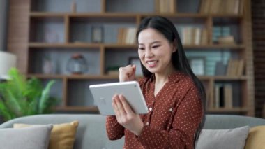 Heyecanlı genç Asyalı kadın spor maçı ya da yarışma izlerken evdeki oturma odasındaki kanepede dijital tablet kullanıyor. Duygusal jestleri olan favori takım için mutlu bayan tezahüratı