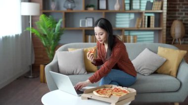 Neşeli, genç Asyalı bir kadın oturma odasında oturmuş pizza yiyor ve dizüstü bilgisayarda video izliyor. Gülümseyen bir dişi hızlı yemek teslimatı konusunda mutludur. Eve yemek götürmek için.