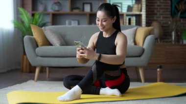 Genç Asyalı sporcu kadın evdeki oturma odasında yerde otururken cep telefonu uygulaması kullanıyor. Spor giyim sektöründe sportif formda bir kadın spor yaparken akıllı telefondan spor uygulamasının keyfini çıkarıyor.