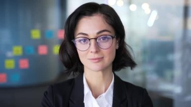 Modern ofiste gözlük takan kendine güvenen bir iş kadını portresi. Takım elbiseli, ciddi bir bayan yöneticinin vesikalığı. Profesyonel kadın kameraya kararlı ve sakin bakıyor.