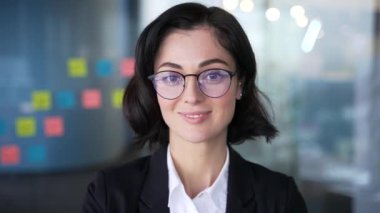 Gözlük takan ve modern ofiste gülümseyen kendine güvenen genç iş kadınının portresi. İş hayatında başarı ve pozitifliği temsil eden profesyonel bir kadın. Takım elbiseli kadın yöneticinin vesikalığı.