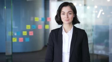 Modern ofiste kollarını kavuşturup gülümseyen kendine güvenen bir iş kadınının portresi. Takım elbiseli genç, pozitif kadın yöneticinin vesikalığı. Profesyonel başarı, girişimcilik ve liderliği temsil eder.